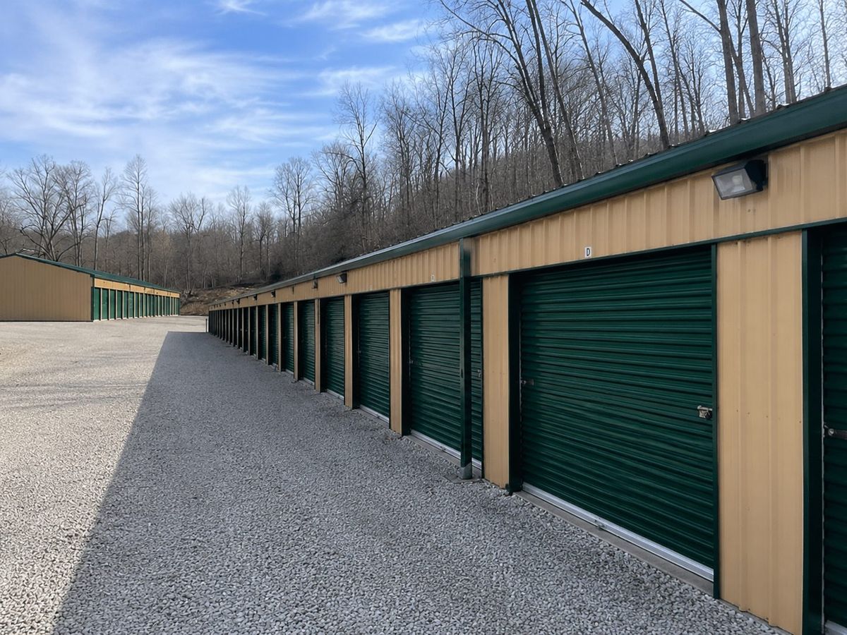 Long side view of a storage unit building showing green roll-up doors, tan metal siding, and a security light on the corner
